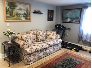 A living room with a floral sofa, a small table with a vase, a treadmill, and a painting on the wall.
