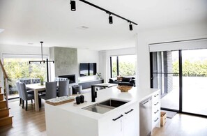A modern kitchen with a central island, dining area with grey chairs, and a large window providing a view of greenery. - Family Getaway with Siete Coramba in Ocean Grove (Ocean Grove)