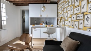 A modern kitchen with white cabinets, a marble countertop, and a small island. There is a dining area with a black sofa and a wooden chair. The wall features framed botanical illustrations.