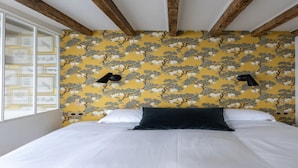 A bedroom with a bed, white bedding, a yellow floral wallpaper, and wooden beams on the ceiling.