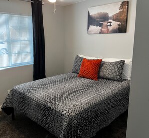 A bedroom with a patterned bedspread, a single red pillow, and a painting of a lakeside scene on the wall.