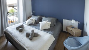 Una cama bien hecha con toallas enrolladas, una mesita de noche con lámpara y una silla en una habitación con paredes azules y suelo de madera.