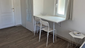 Une petite pièce blanche avec un plancher en bois, un bureau blanc avec une chaise, une fenêtre avec des rideaux et une petite table blanche sur laquelle se trouve un bol.