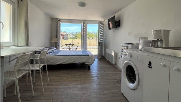 Une pièce de taille modeste comprenant un lit, une petite table, des chaises, un lave-linge et un téléviseur.