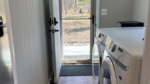 A laundry room with a front door leading to a wooded area, a washing machine, and a drying rack.