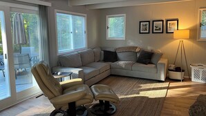 A modern living room with a sectional sofa, a recliner, a small table, and a floor lamp. There are large windows with blinds, a door leading to an outdoor area, and a wall-mounted TV.