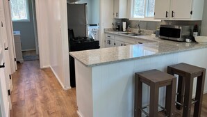 A modern kitchen with white cabinets, a granite countertop, a stainless steel refrigerator, a microwave, and a gas stove. There are two bar stools and a window providing natural light.