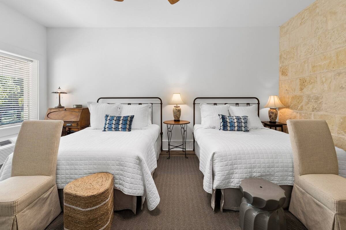 A hotel room with two beds, a ceiling fan, bedside lamps, a small table, and a stone accent wall.