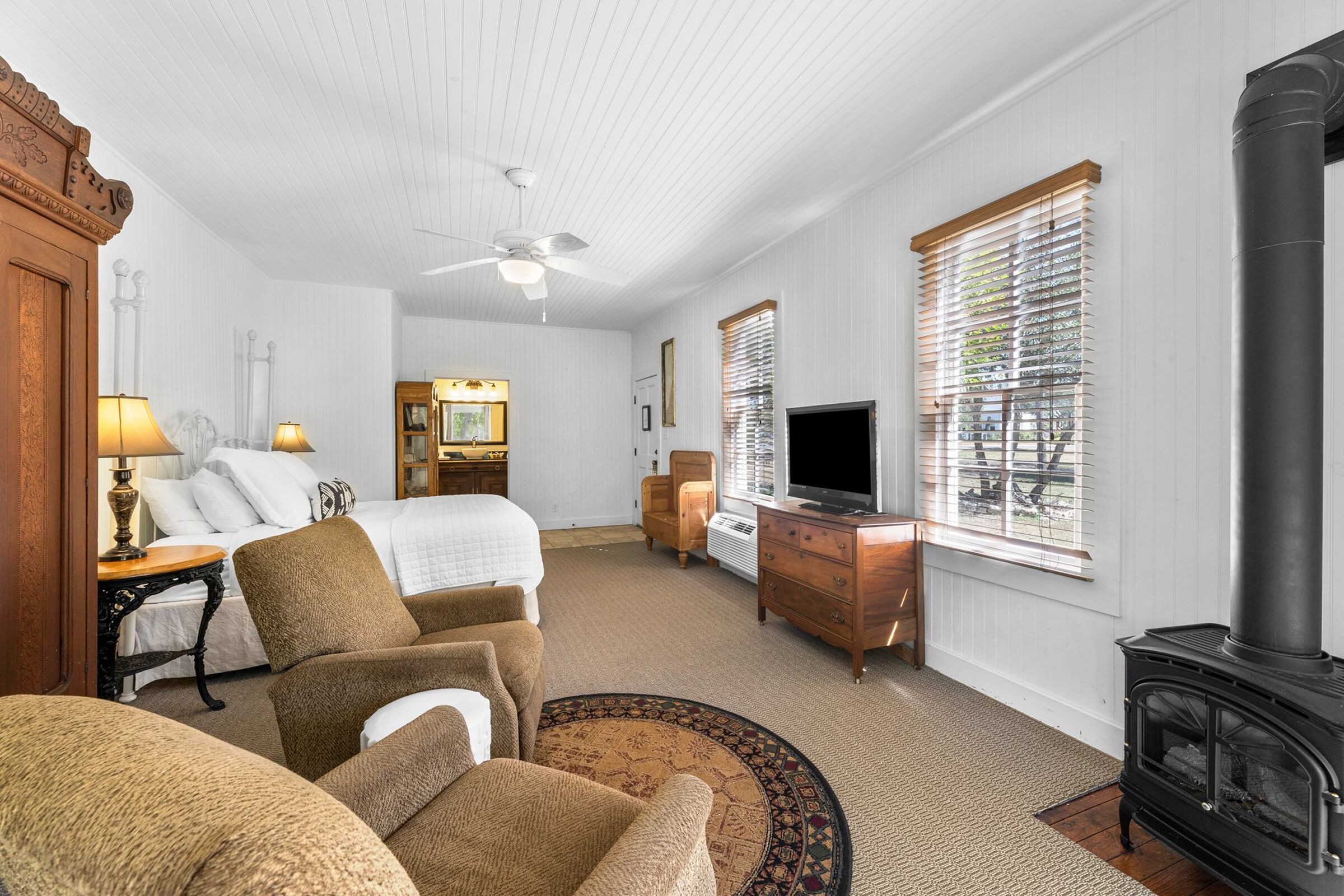 A hotel room with a bed, two armchairs, a TV, a fireplace, a desk, and a window with blinds.
