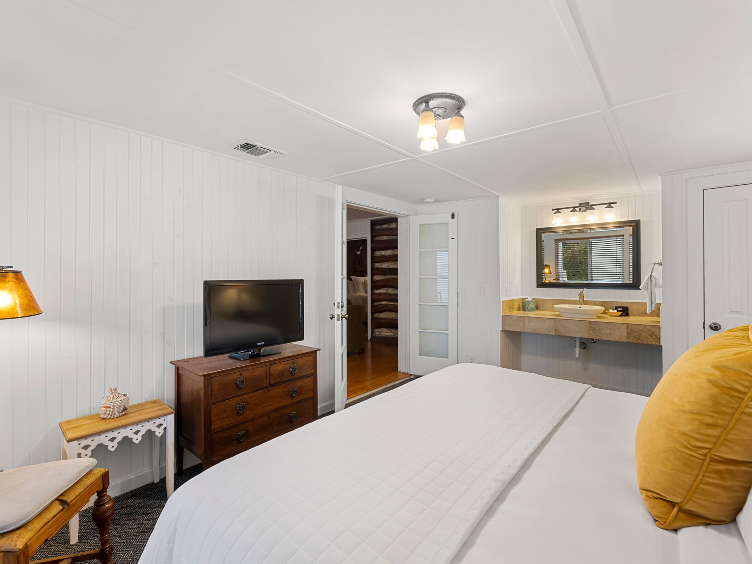 Habitación de hotel con cama, televisor, cómoda de madera, escritorio y baño con lavabo y espejo.