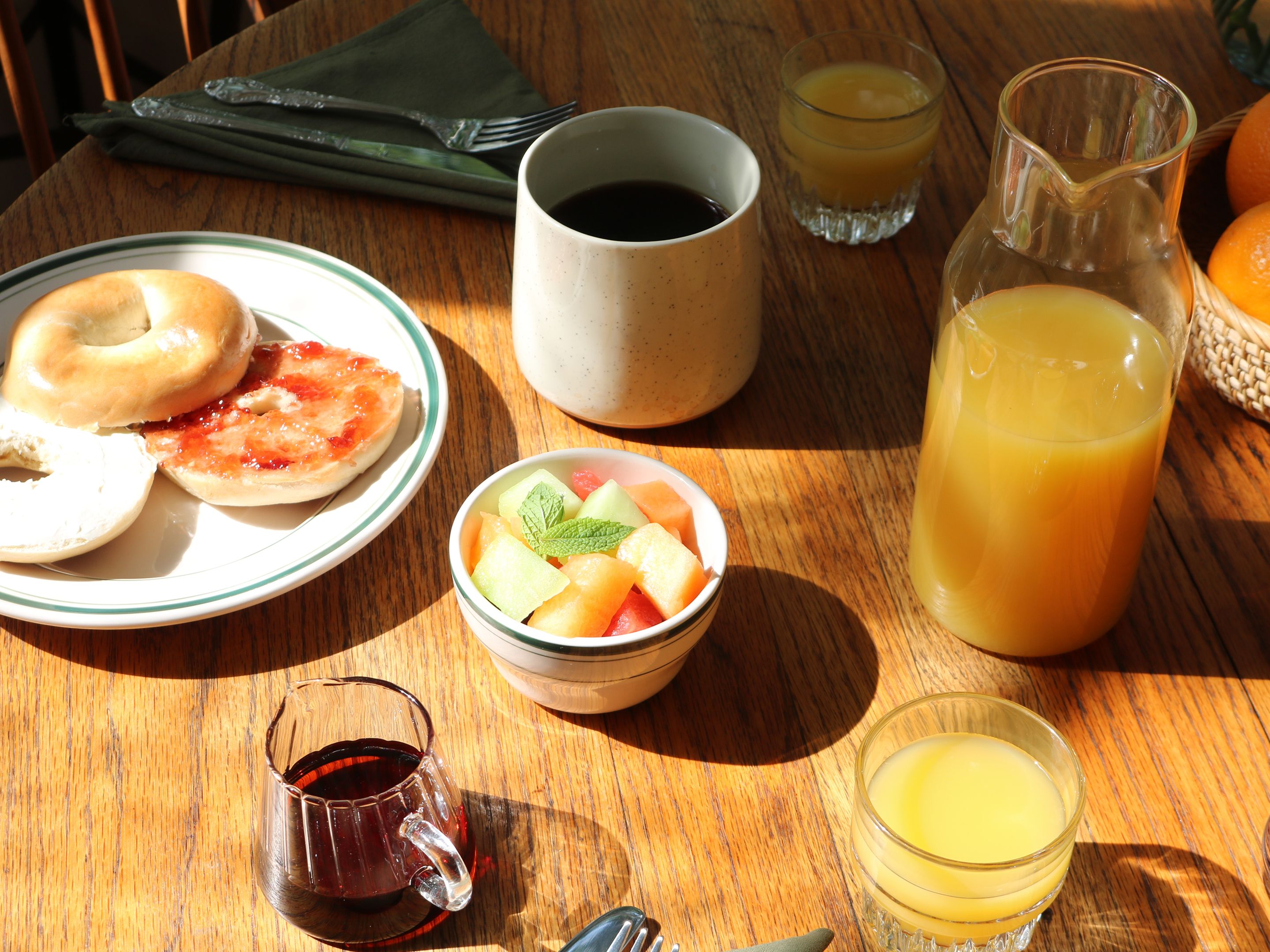 A breakfast setting with a plate of bagels, a bowl of fruit, a cup of coffee, a glass of orange juice, and a pitcher of juice on a wooden table.