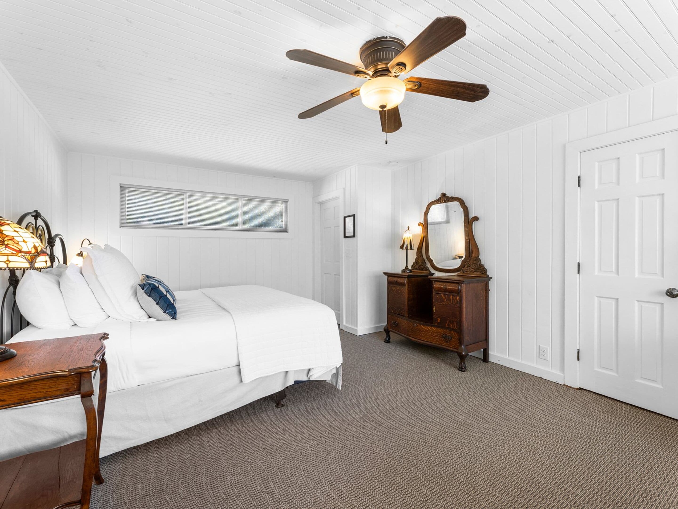 A bedroom with a bed, a dresser, a mirror, a ceiling fan, and a window with blinds.