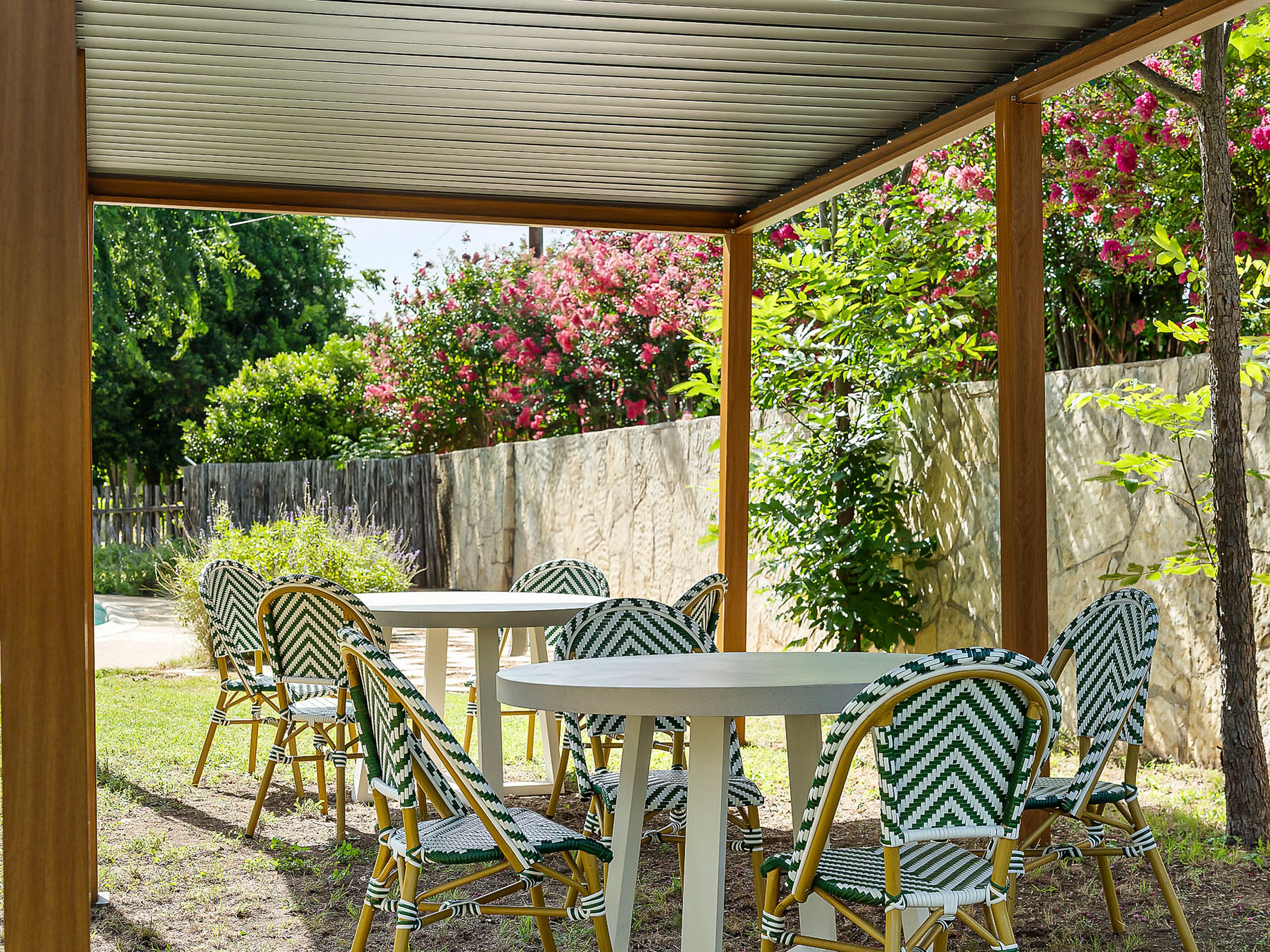 Un patio exterior con sillas de mimbre y una mesa, rodeado por un muro de piedra y abundante vegetación.