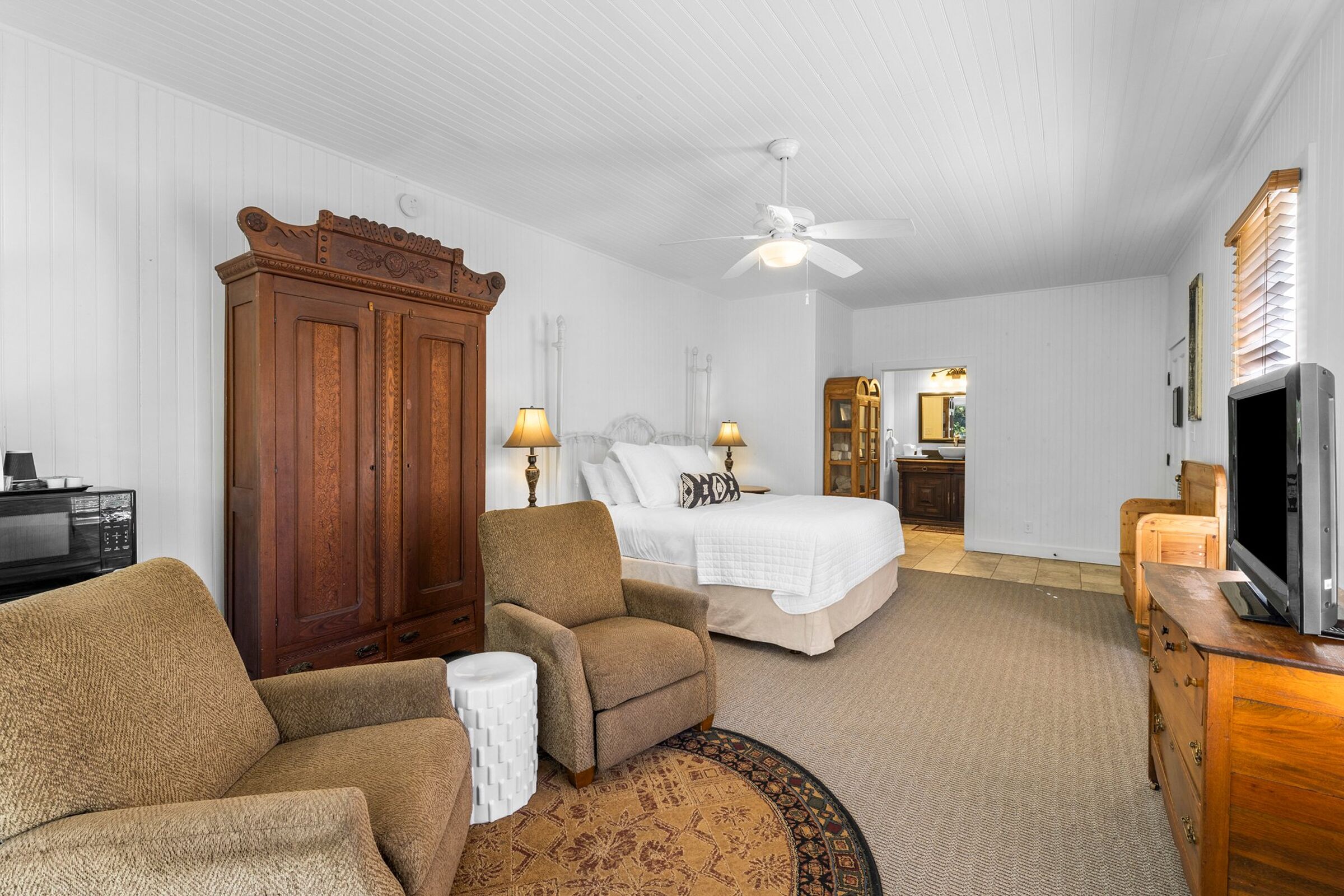 A bedroom with a bed, two chairs, a wooden wardrobe, a television, and a ceiling fan.