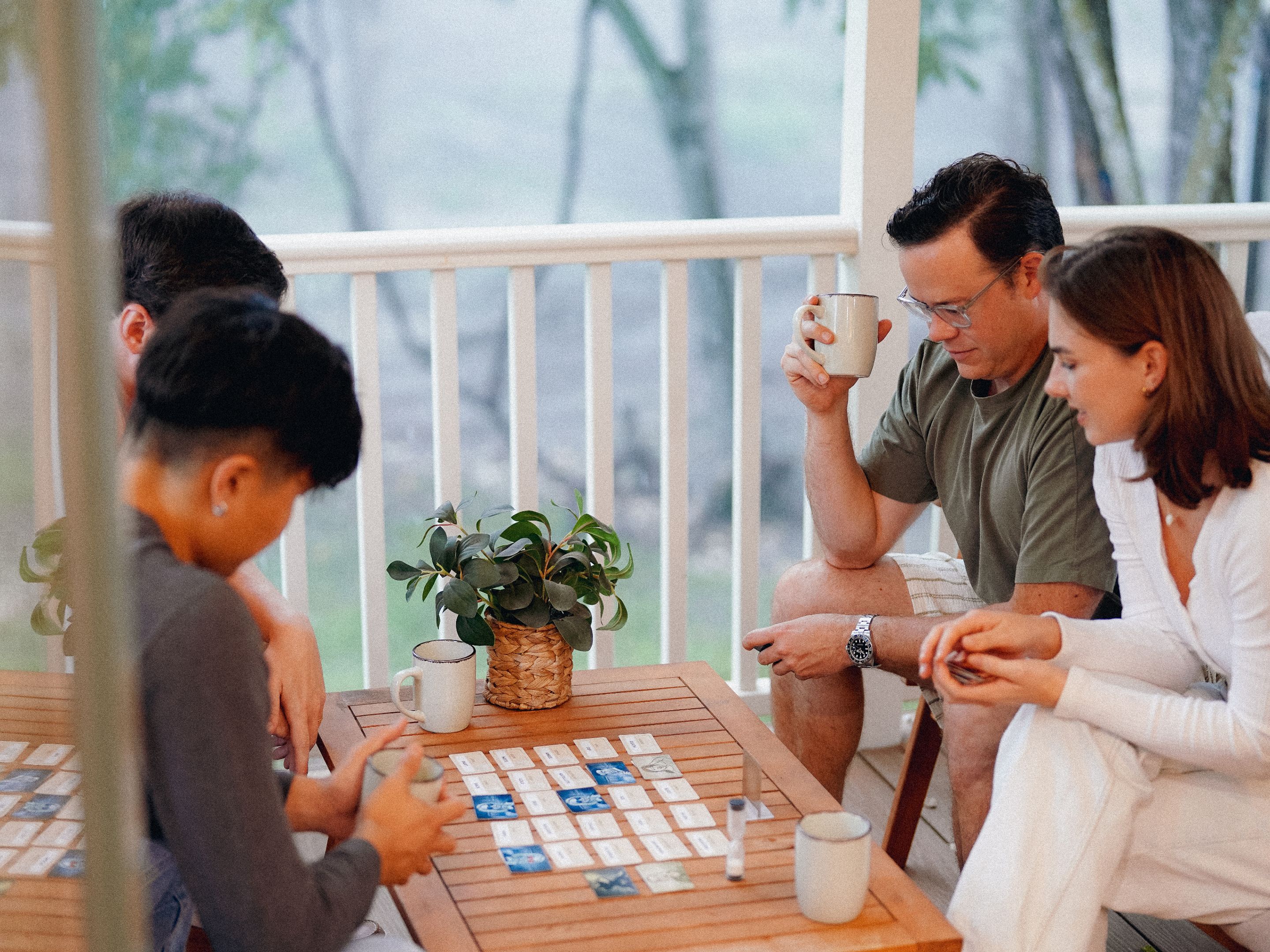 A group of people playing a board game on a wooden table with a potted plant and a white mug on it.