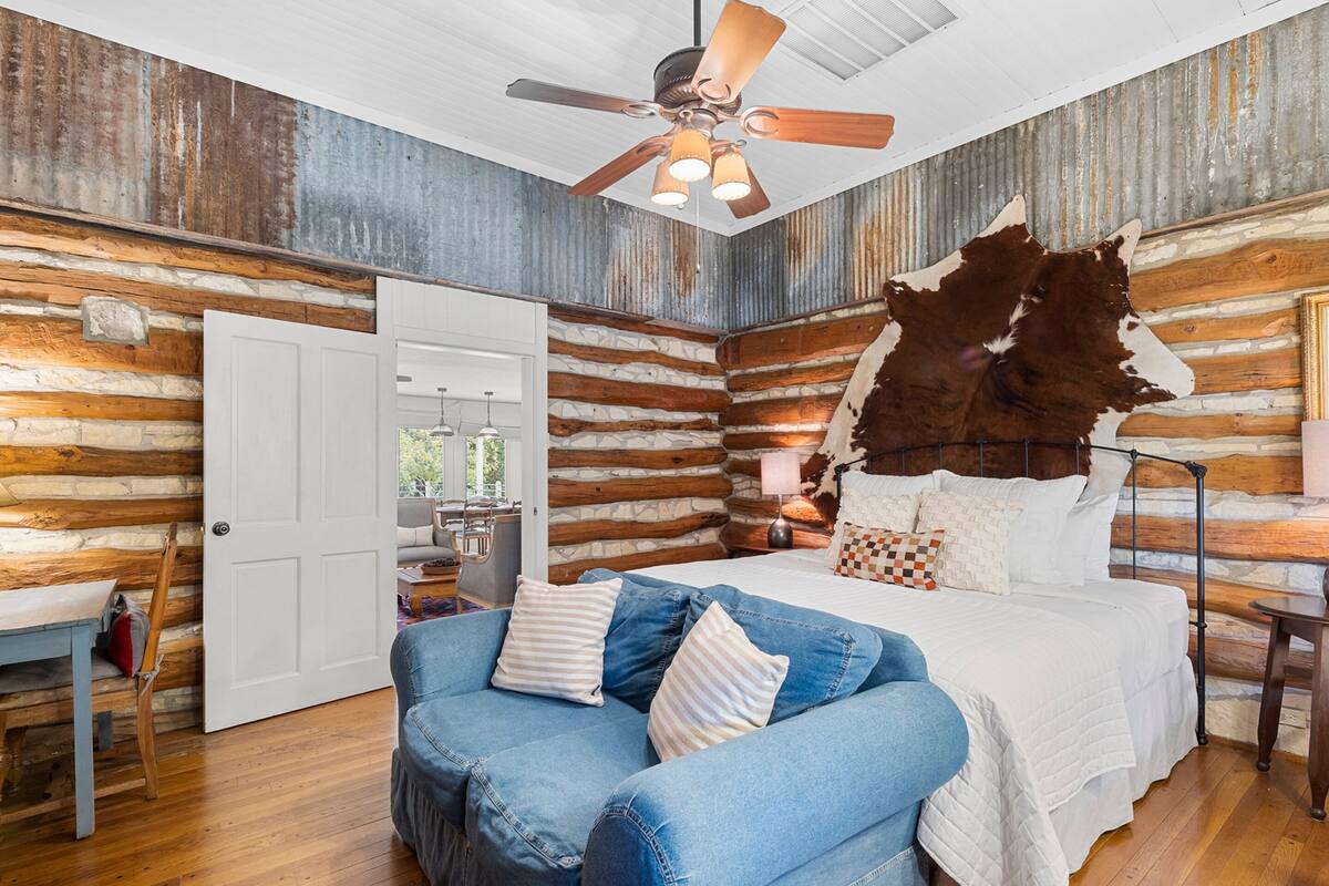 A log cabin-style bedroom with a bed, a blue couch, a ceiling fan, and a rustic wooden wall.