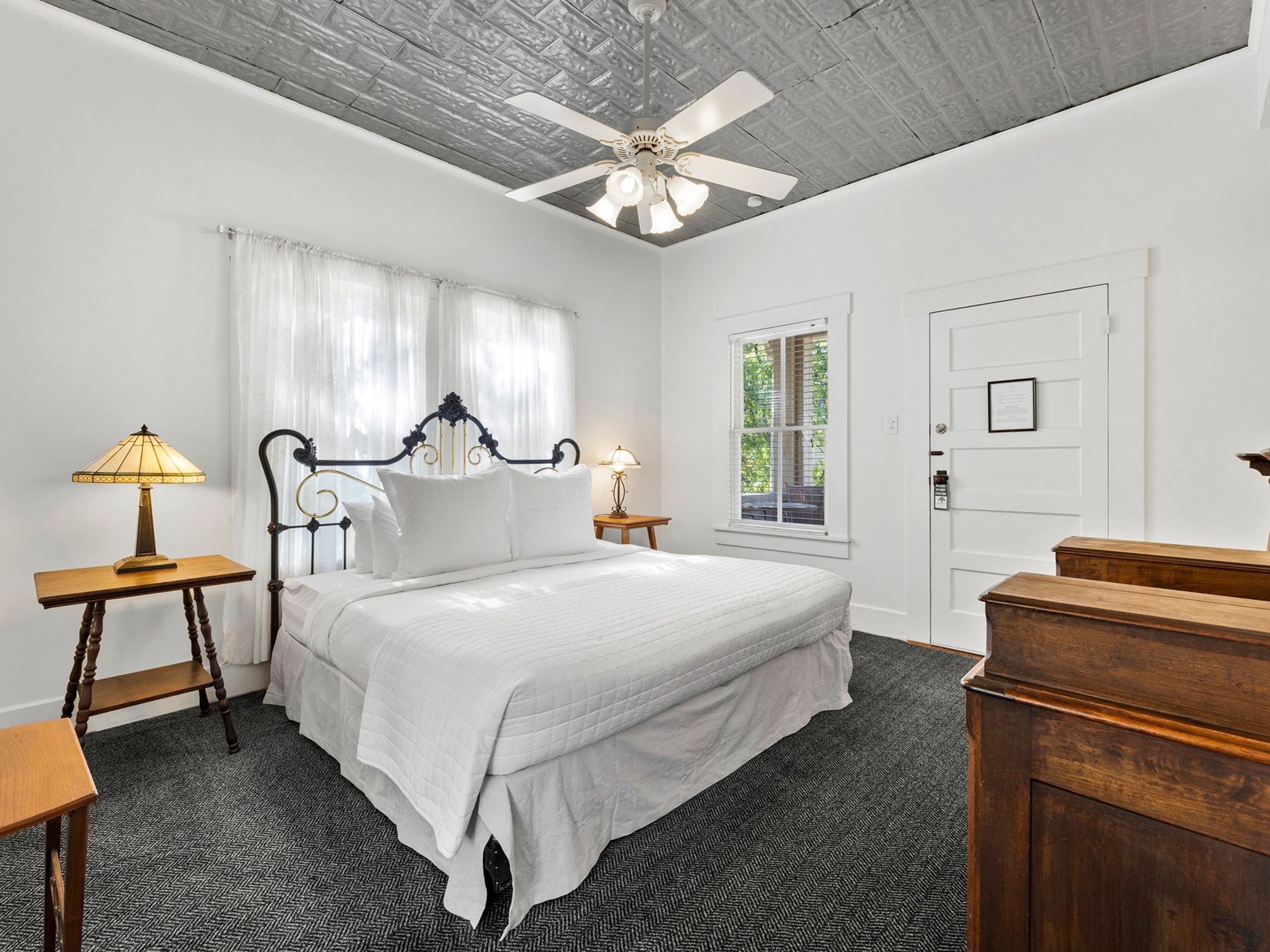 A bedroom with a bed, two bedside tables, a ceiling fan, a window with curtains, and a door.