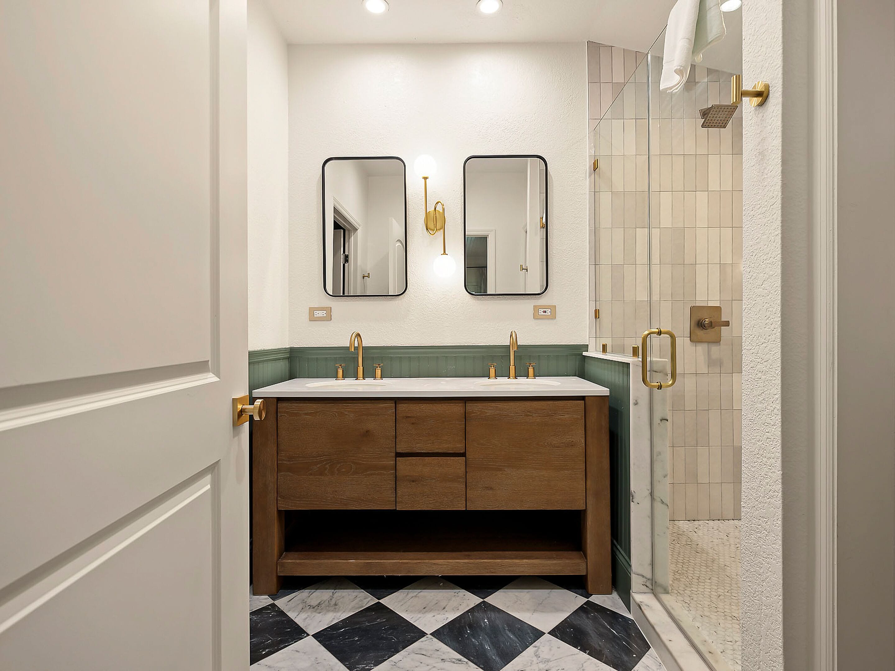 Un baño con tocador doble, dos espejos en la pared, ducha con puerta de cristal y suelo a cuadros.