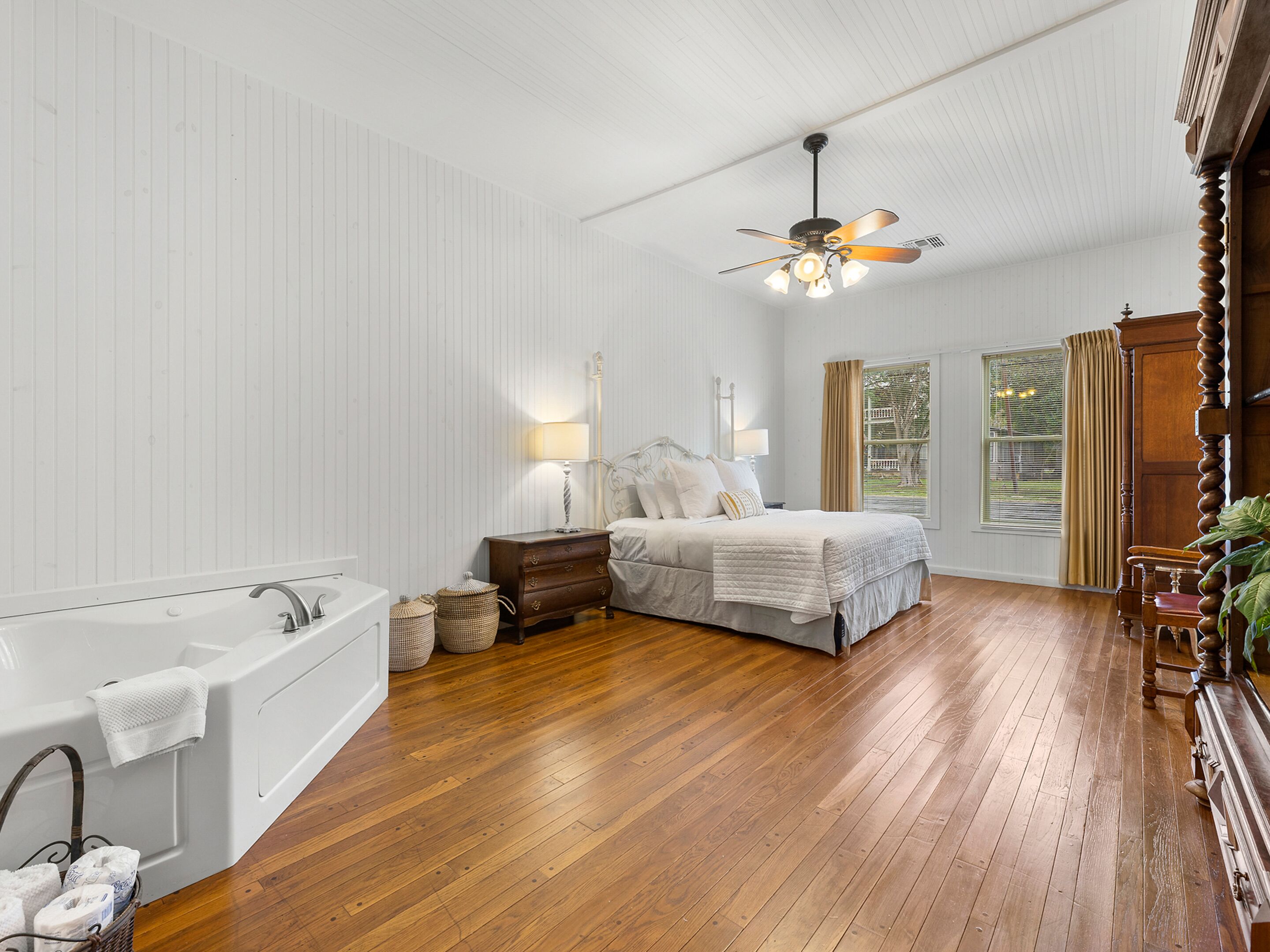A bedroom with a large bed, a nightstand, a lamp, a ceiling fan, a window with curtains, and a wooden floor.