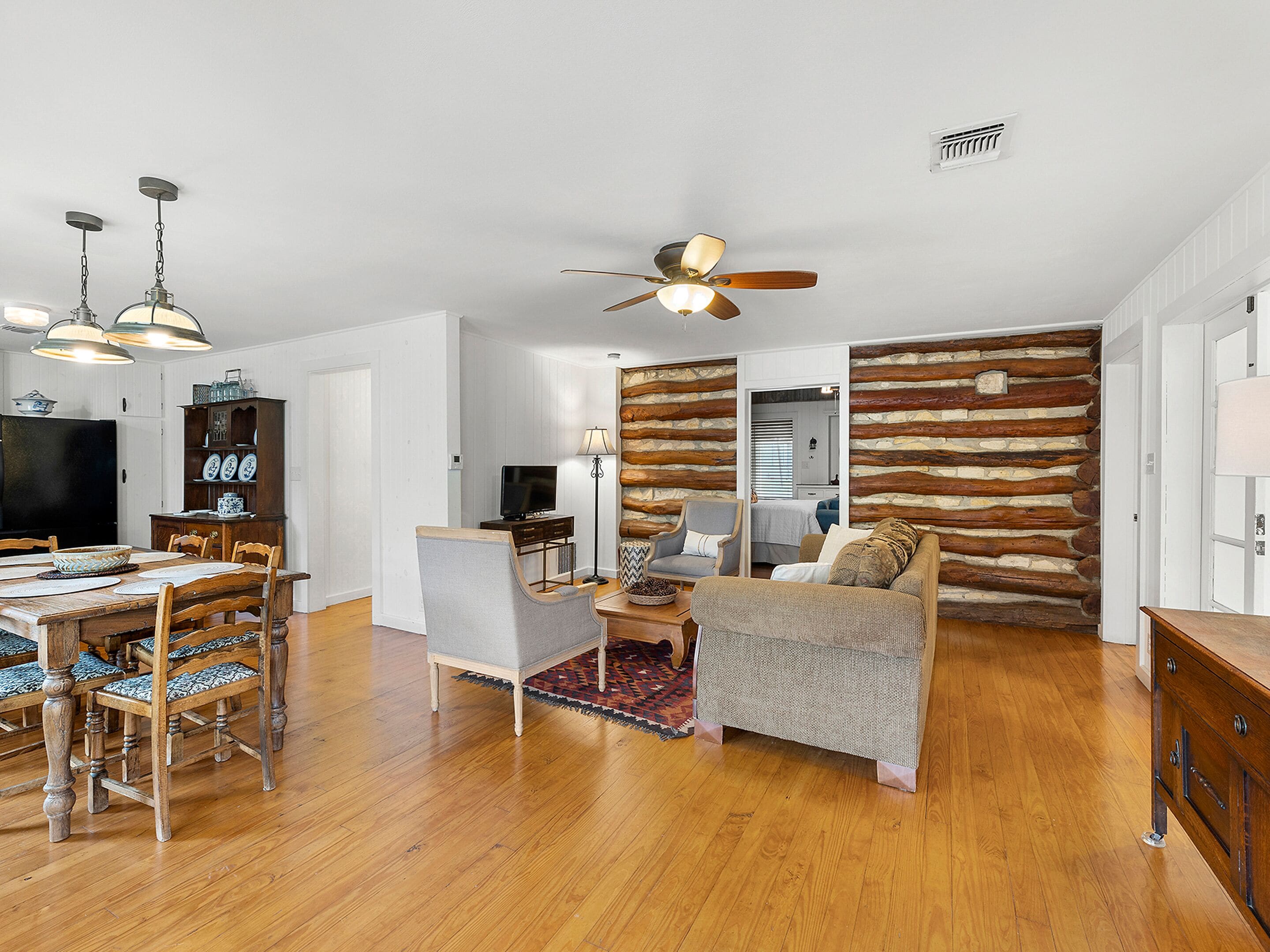 A spacious living area with a wooden floor, a dining table with chairs, a sofa, a TV, and a ceiling fan.
