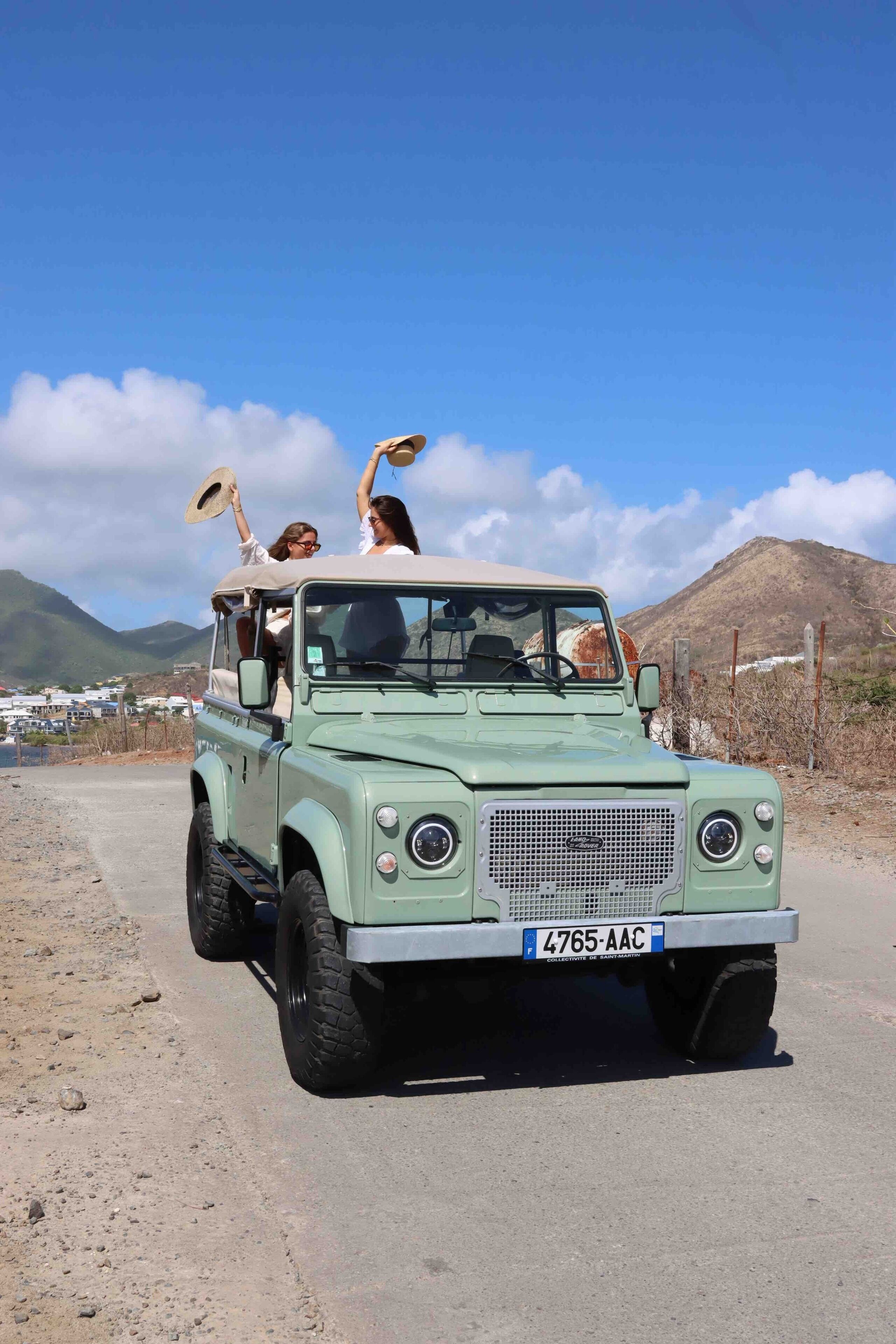 Un véhicule tout-terrain vert transporte deux passagers : l’un tient un chapeau et l’autre un appareil photo. Ils roulent sur un chemin de terre, avec des montagnes en arrière-plan.