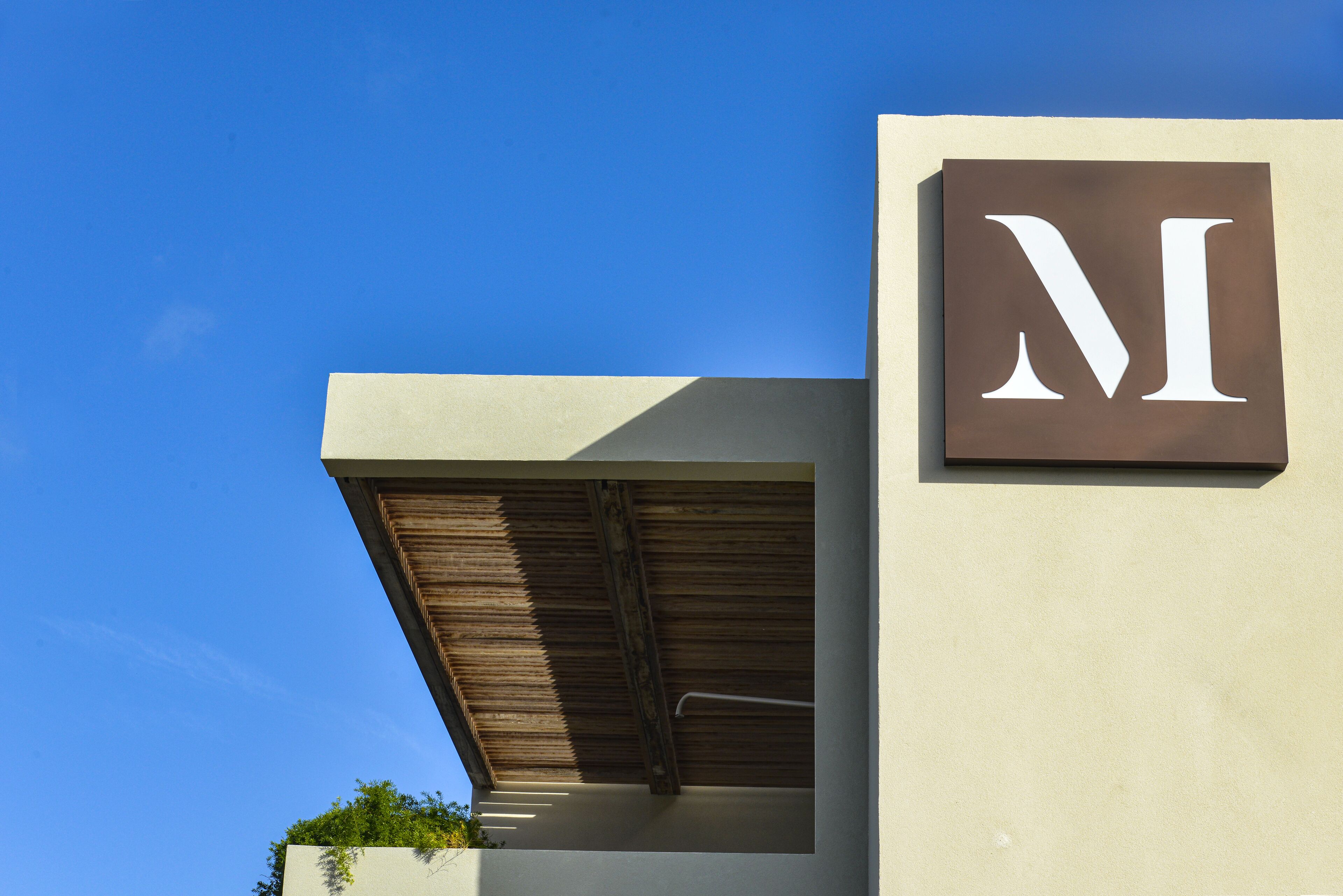 A building with a prominent 'M' sign on a beige wall, a wooden slatted roof, and a clear blue sky in the background.