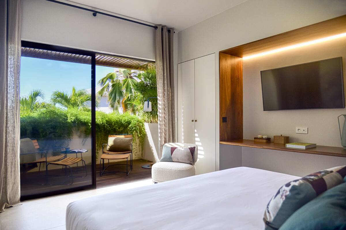 Una camera d'albergo moderna con un letto grande, una TV a parete, un piccolo angolo salotto con una sedia e un tavolino, e una porta scorrevole in vetro che dà su un patio esterno con piante e vista sul cielo.