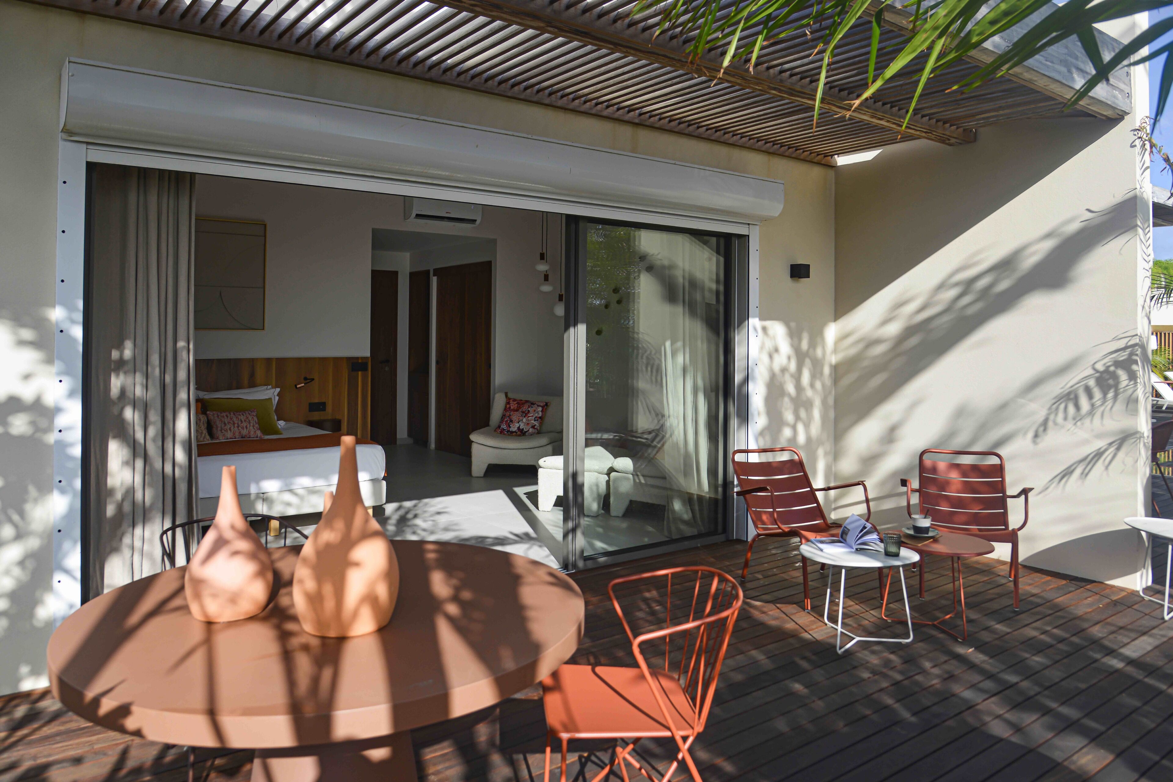 Un área de estar al aire libre con un diseño moderno, una mesa redonda y sillas metálicas, una puerta corredera de vidrio que da a un dormitorio y una terraza de madera.