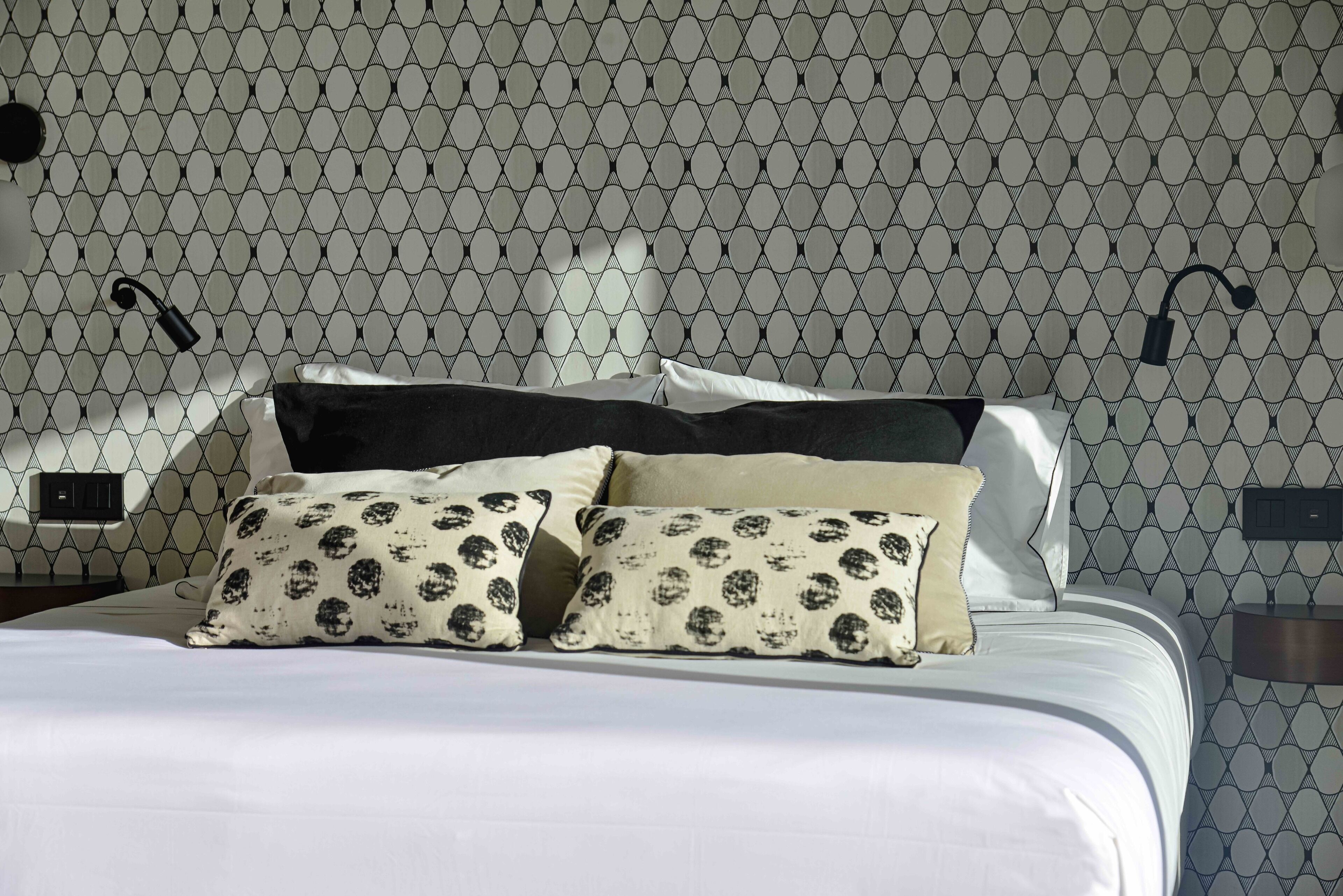 A neatly made bed with patterned pillows, a headboard, and a wall with a geometric pattern.