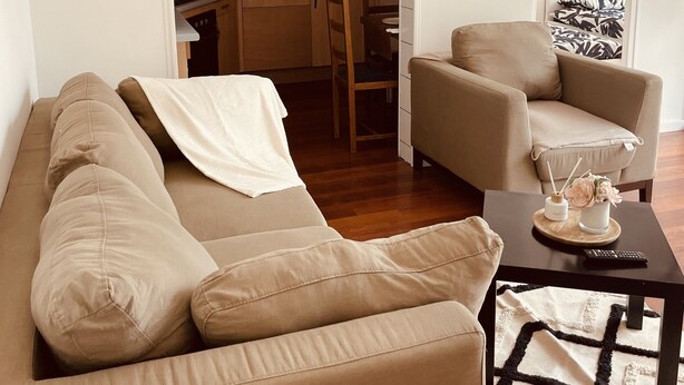 Ein Wohnzimmer mit einer Couch, einem Couchtisch und einem Sessel. Durch einen offenen Durchgang ist eine Küche zu sehen.