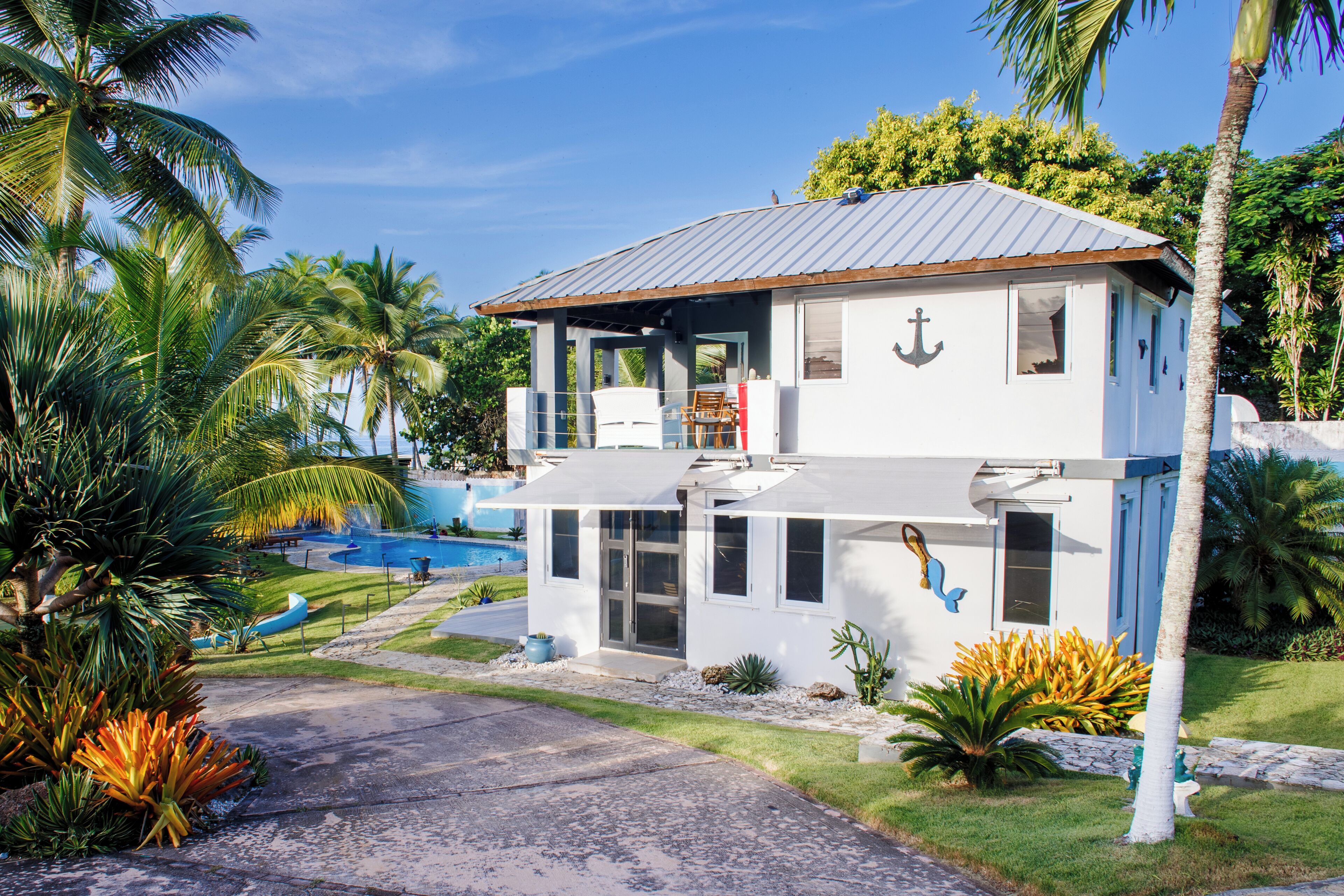 Una casa de dos pisos con balcón, piscina y jardines tropicales.