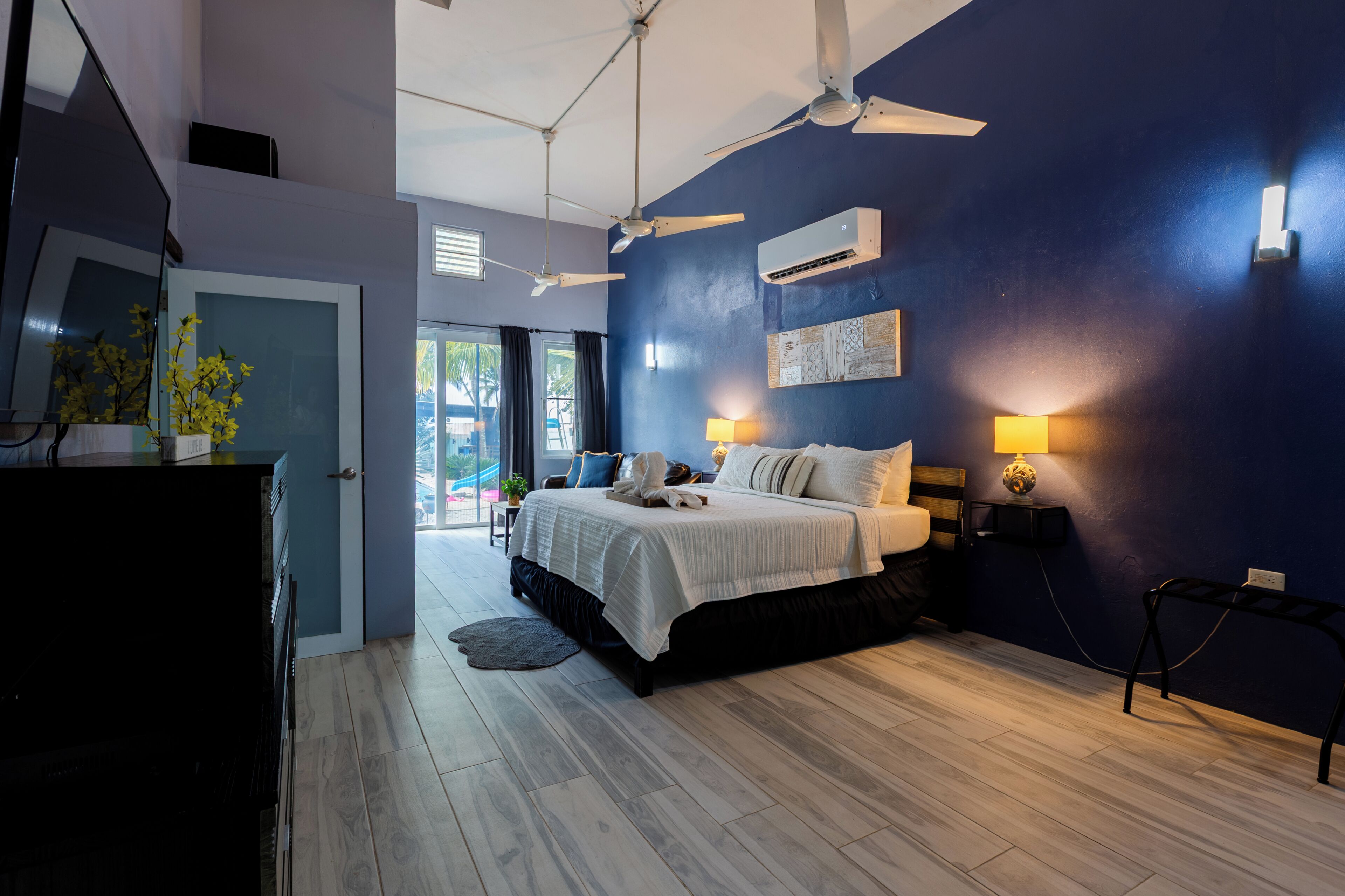 Un dormitorio con cama, ventiladores de techo, aire acondicionado instalado en la pared, lámparas de noche y vistas a través de un ventanal.