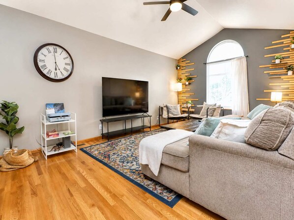 A modern living room with a flat-screen TV, a sofa, a coffee table, a patterned rug, and a wall-mounted clock.