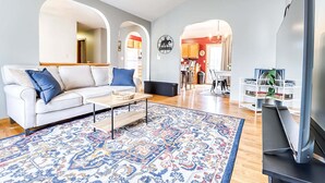 Una sala de estar moderna con un sofá blanco, cojines azules, una mesa de centro y una alfombra estampada. Hay un televisor de pantalla plana sobre un mueble bajo y una estantería con objetos decorativos.