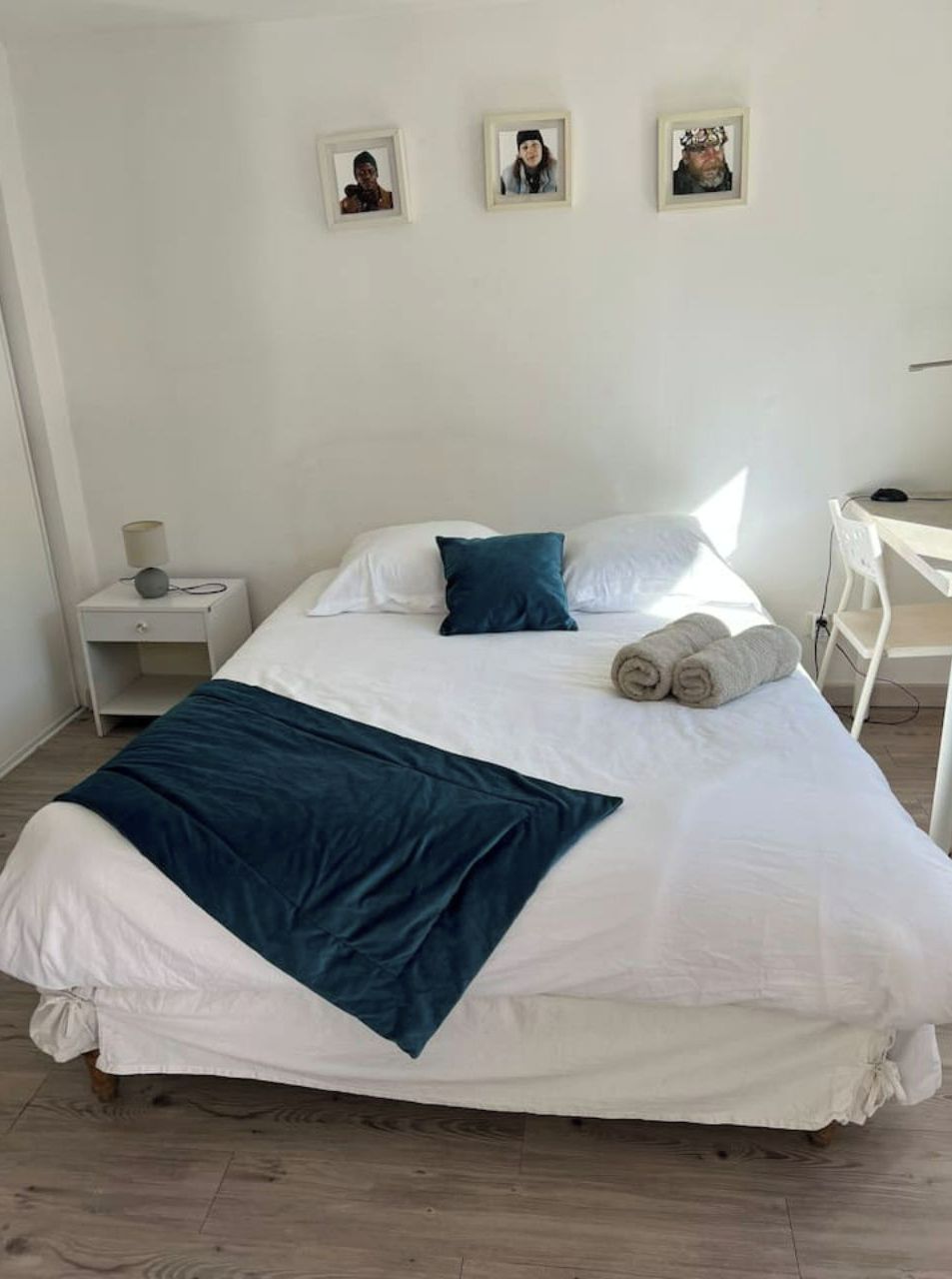 A neatly made bed with white linens and a dark blue throw blanket, a bedside lamp, and a small shelf with a lamp on a white nightstand.
