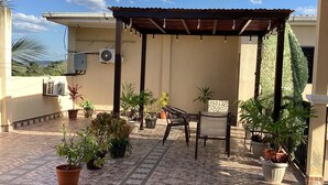 Un patio techado al aire libre con suelo de baldosas, plantas en macetas y una mesa y silla de madera.