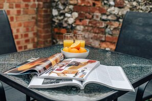 A glass table with a magazine, a bowl of oranges, and glasses of orange juice. - Honeysuckle Terrace - From 125 Per Night (Henley-on-Thames)