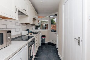 A narrow kitchen with white cabinets, a built-in oven, and a window overlooking a brick wall. - Honeysuckle Terrace - From 125 Per Night (Henley-on-Thames)