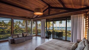 Une chambre spacieuse avec un grand lit, un canapé et un balcon offrant une vue sur les palmiers et l’océan.