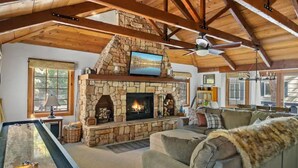 A cozy living room with a stone fireplace, wooden beams, a ceiling fan, and a pool table.