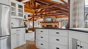 A kitchen with white cabinets, a stone fireplace, and exposed wooden beams.