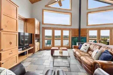 A spacious living room with a large wooden TV stand, a leather sofa with cushions, a glass coffee table, and a view of the outdoors through large windows.