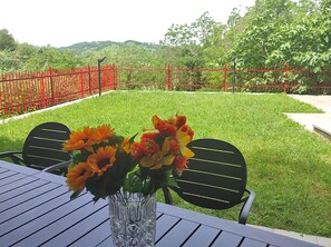 A table with a vase of flowers and two chairs, set on a lawn with a red fence and greenery in the background.
