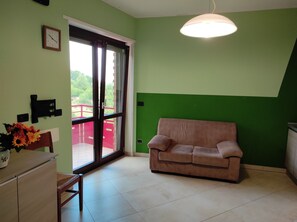 A room with a green wall, a brown sofa, a wooden cabinet, and a window with a view of a red railing and greenery outside.