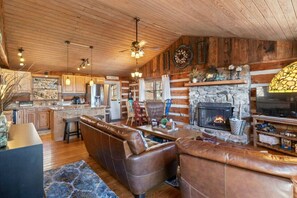 A rustic cabin interior with a stone fireplace, leather seating, wooden floors, and a kitchen area. - Wolf Laurel-Authentic Cabin-Mountain Views-Ski Hatley Pointe-Hiking-A/T-Big Bald (Mars Hill)