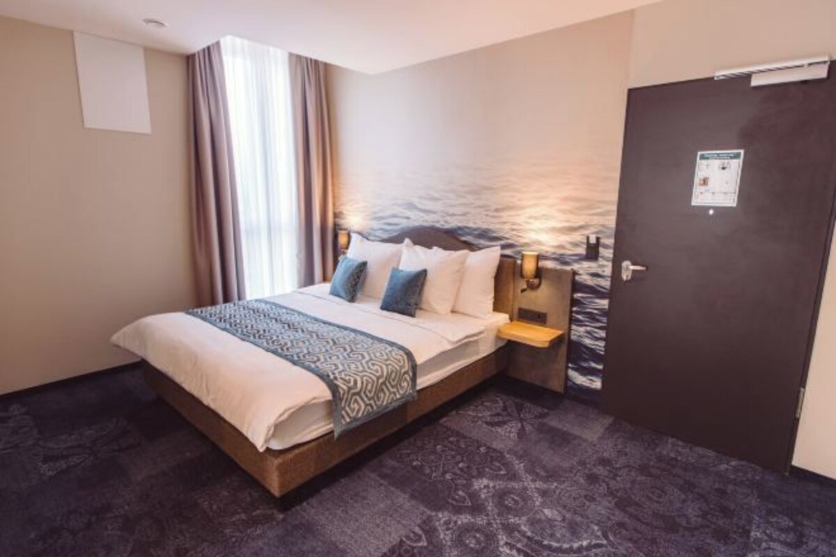 A hotel room with a bed, a nightstand, a door, and a view of the sea through the window.