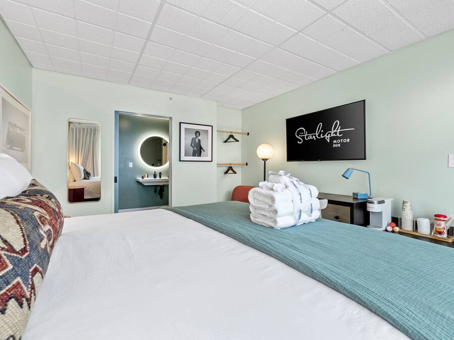 A hotel room with a bed, a desk, a mirror, a wall-mounted TV, and a door leading to a bathroom.