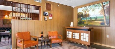 A room with wooden panel walls, a checkered rug, two leather chairs, a small table with a lamp, and a painting of a tropical landscape.