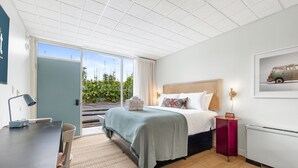 A hotel room with a bed, a desk, a chair, a radiator, and a window with a view of greenery.