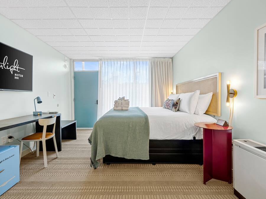 A hotel room with a bed, a desk with a chair, a television, a heater, and a window with curtains.